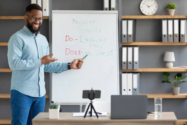 Χαρούμενη χιλιετή μαύρος δάσκαλος με γυαλιά κοιτάζει τη συσκευή, δείχνει στον πίνακα με αγγλικούς κανόνες — Φωτογραφία Αρχείου