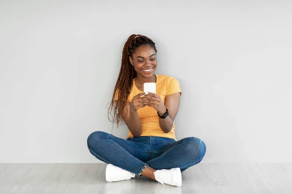 Mujer negra usando teléfono móvil, video chat en la web, trabajando o estudiando en línea, sentada en el suelo contra la pared gris — Foto de Stock