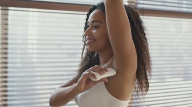 Kişisel hijyen konsepti. Genç Afrikalı Amerikalı kadın deodorant sürüyor, banyoda gülümsüyor, çekim takibi yapıyor.