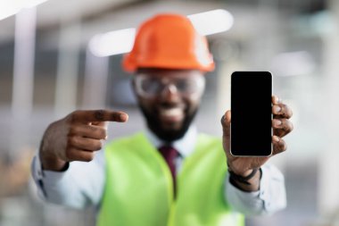 Gülümseyen Afro-Amerikan inşaat mühendisi boş ekranlı akıllı telefon gösteriyor.