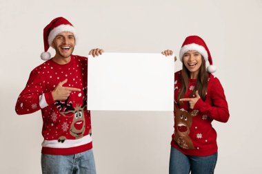 Tatil reklamı. Neşeli eşler Noel Baba şapkaları takıyor Boş Beyaz Pankartı işaret ediyorlar