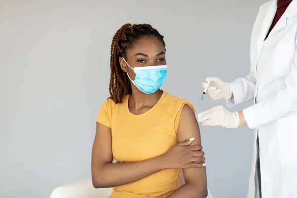 Female doctor making injection of anti-covid vaccine to young black woman in medical mask, grey studio background — 图库照片