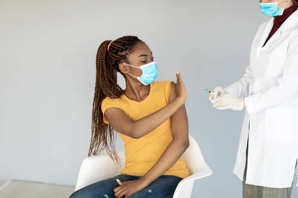 Jeune femme noire en masque facial jetable empêchant le médecin de faire une injection de vaccin contre le coronavirus sur fond gris — Photo