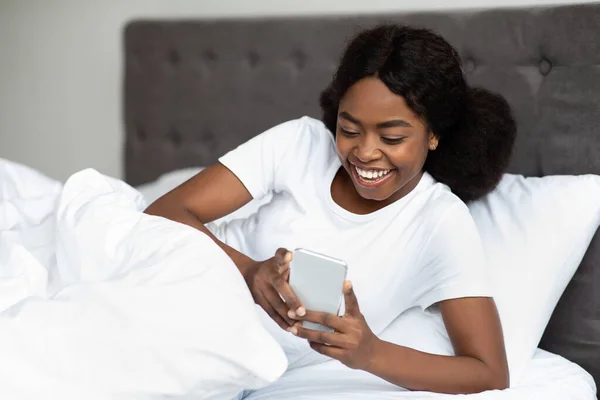 Улыбающаяся черная леди с помощью мобильного телефона, когда лежит в постели — стоковое фото