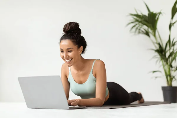 Mujer joven sonriente en ropa deportiva acostado en la estera de fitness y el uso de ordenador portátil —  Fotos de Stock