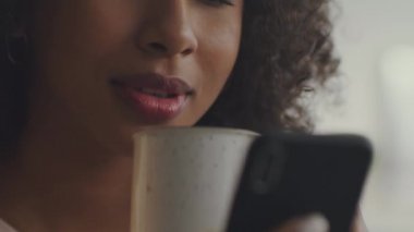 Modern sabah alışkanlıkları. Genç Afro-Amerikan kadınının sıcak kahve içerken ve akıllı telefondan haber okurken yakın plan çekimleri.