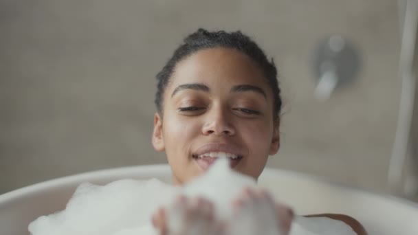 Jong gelukkig Afrikaans amerikaans vrouw ontspannen in schuimig warm bad blazen schuim op camera en glimlachen, close-up portret — Stockvideo