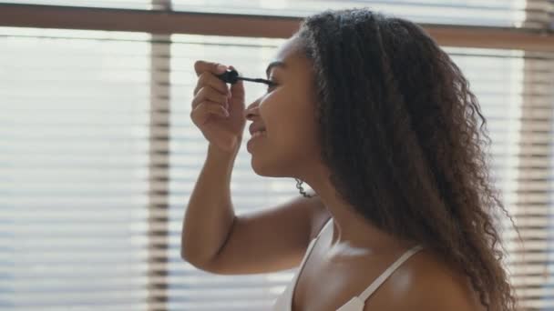 Maquillaje todos los días. Perfil retrato de la joven dama afroamericana feliz aplicando rimel en las pestañas, seguimiento de disparo — Vídeo de stock