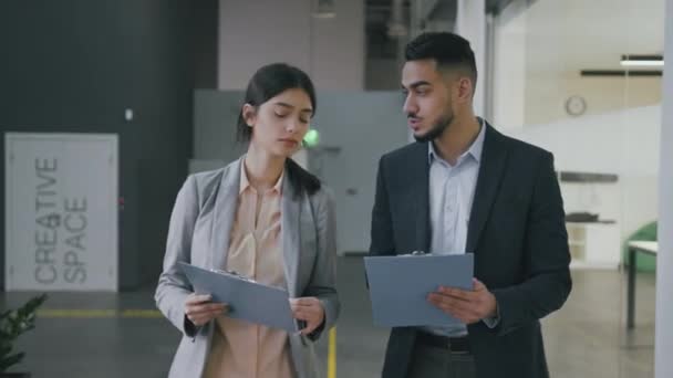 Two young diverse colleagues walking along modern office building with documents and discussing project — Stockvideo