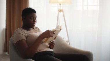 Büyük boy Afrikalı Kadın Evde Sandalyede Oturarak Kitap Okuyor