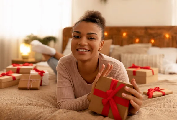 침대에 누워 크리스마스 선물 상자를 손에 쥐고 카메라 앞에서 미소짓는 행복 한 젊은 아프리카계 미국 여성 — 스톡 사진
