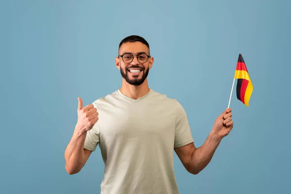 Felice ragazzo arabo mostrando pollice in su e la bandiera della Germania, in posa su sfondo blu studio. Istruzione in Germania — Foto Stock