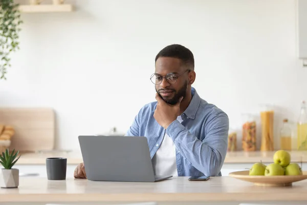 Серьезный африканский бородатый бизнесмен тысячелетия в очках работает на ноутбуке и обдумывает решение проблем — стоковое фото