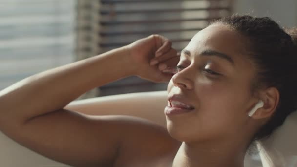 Jeune femme afro-américaine insouciante écoutant de la musique et chantant, relaxant dans la salle de bain avec écouteurs sans fil — Video