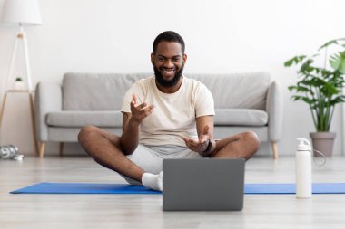 Beyaz elbiseli, gülümseyen siyah sakallı koç mindere oturur, bilgisayara bakar, iletişim kurar ve jestler yapar.