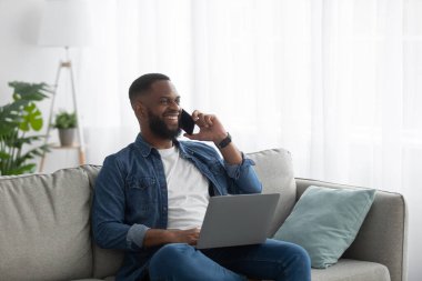 Mutlu Afro-Amerikan sakallı adam bilgisayarda yazı yazıyor ve oturma odasındaki kanepede telefonla arıyor.