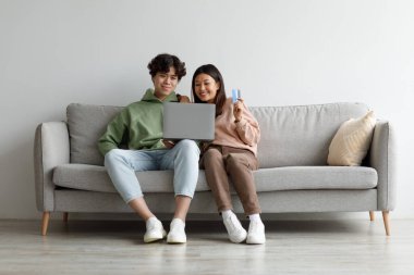 İnternetten alışveriş. Dizüstü bilgisayarı ve kredi kartı olan genç bir Asyalı çift kanepede oturmuş evdeki web mağazasından mal alıyorlar.