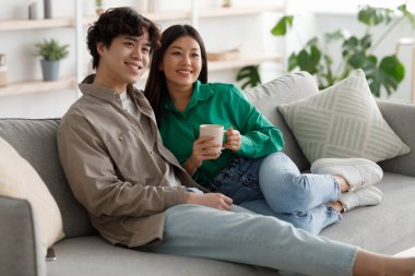 Mutlu hafta sonları. Sevecen genç Asyalı çift kahve içiyor ve evdeki rahat kanepede sarılıyorlar.