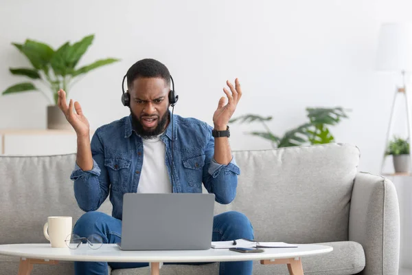 Sorprendido disgustado joven barbudo negro hombre en los auriculares levanta las manos, hablar por ordenador portátil y gesto — Foto de Stock