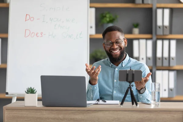 Ευτυχής νεαρός μαύρος δάσκαλος, δάσκαλος σε γυαλιά κοιτάζει φορητό υπολογιστή και τηλέφωνο κάμερα στο εσωτερικό του γραφείου στο σπίτι με μαυροπίνακα — Φωτογραφία Αρχείου