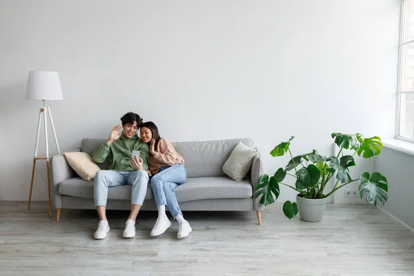 Alegre jovem casal asiático acenando para câmera do smartphone, conversando online, tendo videoconferência no sofá em casa — Fotografia de Stock