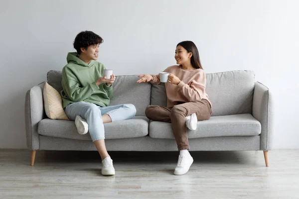 Νεαρός Ασιάτης άνδρας και γυναίκα πίνει καφέ και μιλάει ενώ κάθεται στον καναπέ στο σπίτι — Φωτογραφία Αρχείου