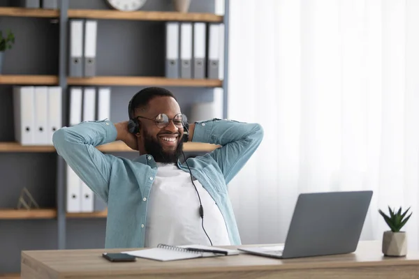Gelukkig jong zwart baardman in bril en koptelefoon verheugt zich in succesvolle deal, ontspannen en kijkt naar laptop — Stockfoto