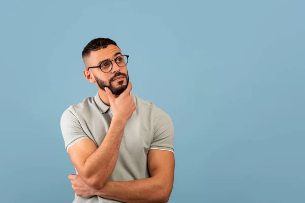 Pensive middle eastern guy thinking and touching chin, looking aside at free space, posing over blue background — Stockfoto