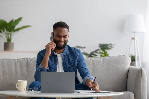 Χαμογελώντας νεαρό αφροαμερικανό γενειοφόρο αρσενικό δακτυλογράφηση σε φορητό υπολογιστή, μιλώντας στο τηλέφωνο με τον πελάτη — Φωτογραφία Αρχείου