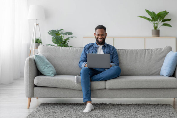 Happy young black bearded male chatting in social network, businessman work on laptop on sofa