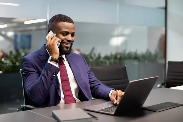 Homme d'affaires afro-américain prospère travaillant sur un ordinateur portable, ayant une conversation téléphonique — Photo