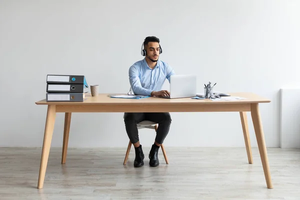 Uomo arabo che utilizza il computer portatile indossando auricolare seduto alla scrivania — Foto Stock