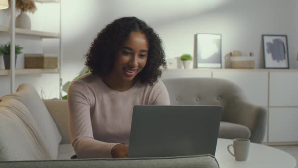 Joven mujer afroamericana positiva freelancer escribiendo en el ordenador portátil, charlando con los clientes y disfrutando de trato en casa — Vídeo de stock