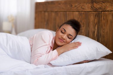 Sağlıklı uyku konsepti. Barışçıl Afrikalı Amerikalı kadın evinde yatağında uyuyor, beyaz, rahat yatak odasında dinleniyor.