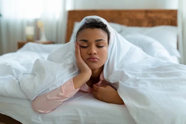 Noche sin dormir, problemas e insomnio. Joven mujer afroamericana con los ojos cerrados acostada debajo de una manta en la cama —  Fotos de Stock