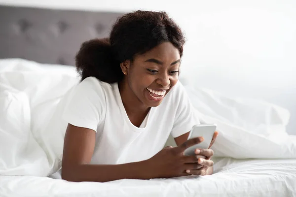 Emocional mujer afroamericana que se enfría en la cama con teléfono inteligente —  Fotos de Stock