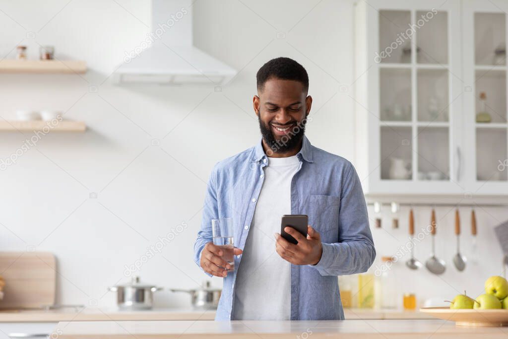 Happy young black man with glass of clear water in hand in minimalist kitchen interior, chatting on phone