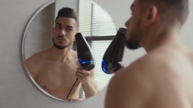 Genç yakışıklı Arap adam banyoda saçını kurutuyor, dışarı çıkmadan önce saçlarını tarıyor, banyoda aynaya bakıyor.