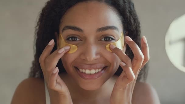 Зеркальный портрет молодой позитивной африканской женщины, надевающей коллагеновые пластыри под глазами в ванной комнате — стоковое видео