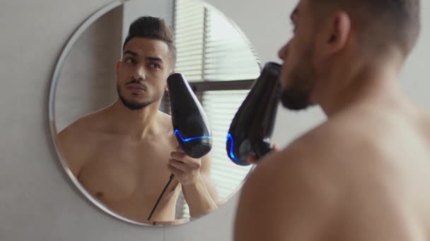 Genç yakışıklı Arap adam banyoda saçını kurutuyor, dışarı çıkmadan önce saçlarını tarıyor, banyoda aynaya bakıyor. — Stok video