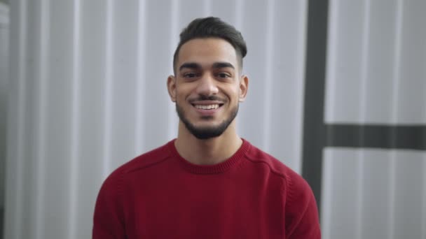 Positif bien entretenu jeune gars du Moyen-Orient portant pull rouge souriant à la caméra, posant sur fond de mur gris — Video