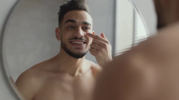 Jovem atraente árabe cara aplicando creme nas bochechas e sorrindo para reflexão, preocupando-se com sua aparência no banheiro — Vídeo de Stock
