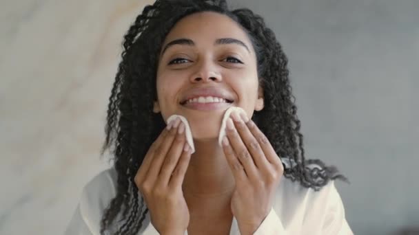 Hozd rendbe magad. Boldog afro-amerikai nő tisztítja arcát micelláris vízzel és két pamut párna, tükör pov — Stock videók