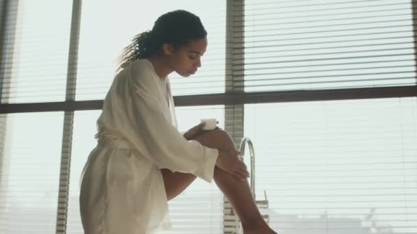 Körperpflege nach dem Duschen. Junge Afroamerikanerin trägt Bademantel und trägt nährende Körpercreme auf die Beine — Stockvideo