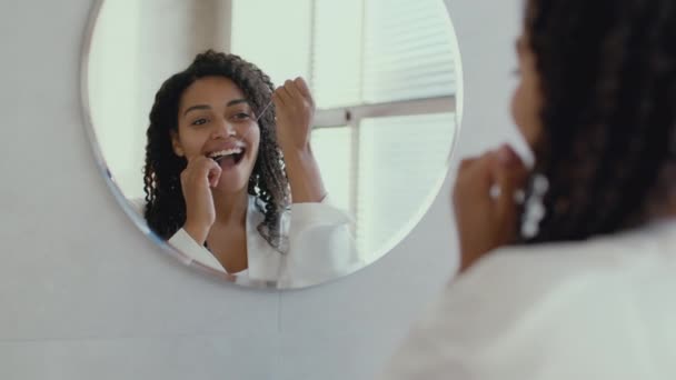 Young positive african american lady in bathrobe flossing her teeth, looking at mirror at bathroom in morning, zoom in — стоковое видео