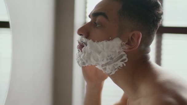 Morgonmännens rutin. Närbild profil porträtt av unga Mellanöstern kille rakning ansikte, titta på spegel på badrummet — Stockvideo