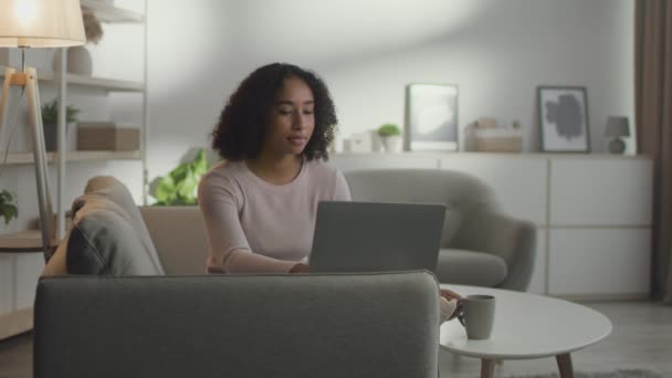 Mladý pozitivní africký Američan žena na volné noze pracuje vzdáleně na notebooku, těší horké kávy — Stock video