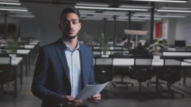 Resmi takım elbiseli genç Arap işadamı kameraya gülümsüyor, modern ofis içinde poz veriyor, izleme görüntüsü, serbest alan