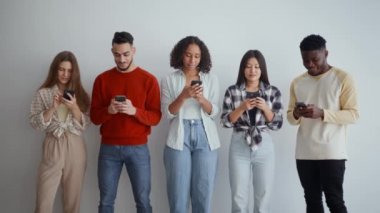 Modern internet iletişimi. Akıllı telefonlarda yazan, internette gezinen, birbirlerini görmezden gelen, beyaz duvara yaslanmış, ağır çekimde çalışan genç kadın ve erkeklerden oluşan bir grup.