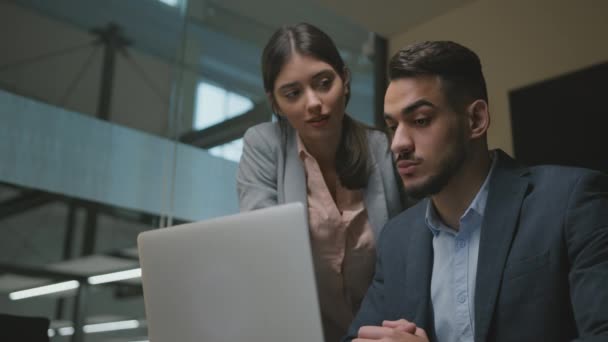 Jovem do Oriente Médio recém-chegado estudo para trabalhar no laptop no novo emprego, jovem mulher ajudando-o no escritório, rastreamento tiro — Vídeo de Stock